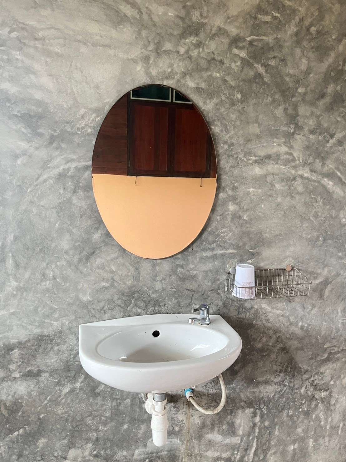 A wall-mounted sink is featured, displaying a simple design with a rounded mirror positioned above. Textured gray walls provide a rustic backdrop, while a small wire basket is attached beside the sink, presumably for toiletries.