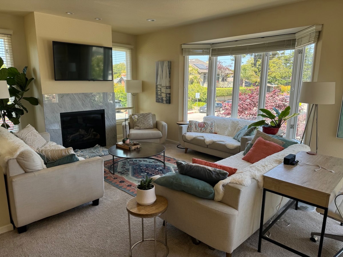 A welcoming living area is featured, with a cozy seating arrangement that includes a plush sofa and comfortable chairs. A minimalist coffee table and a light wooden desk are present. Natural light fills the space through large bay windows, offering views of the outdoor greenery.