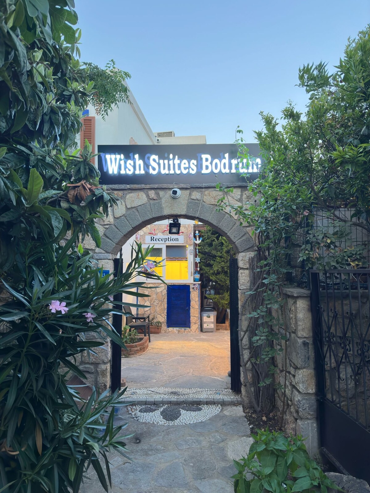 The entrance to Wish Suites Bodrum is framed by lush green plants, leading to a stone archway. A sign above displays the name, while a reception area is visible in the background. The pathway is lined with decorative tiles, creating an inviting atmosphere.