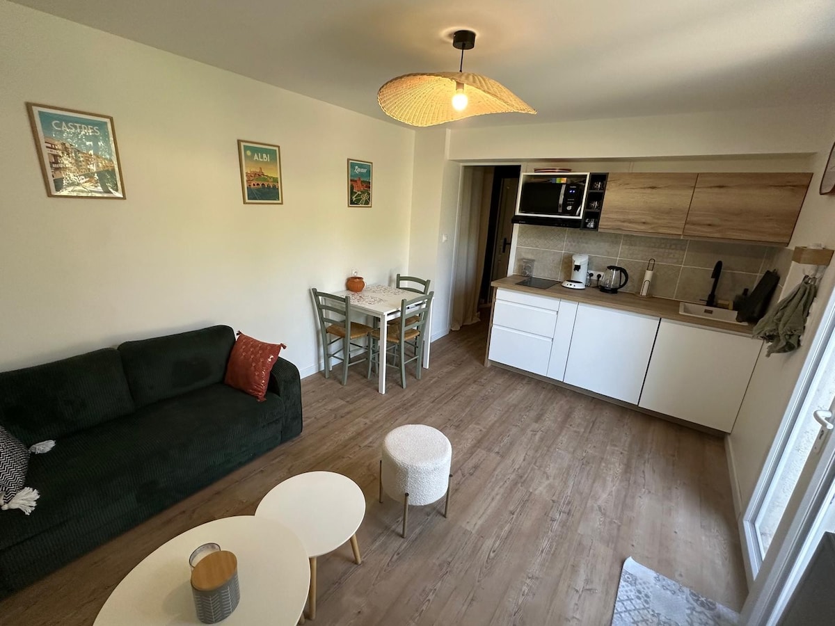 An open living area features a dark green sofa with a decorative orange pillow, accompanied by a light wooden dining table and four chairs. A kitchen area is in the background, offering modern appliances and wooden cabinetry. Natural light streams in through a nearby window.