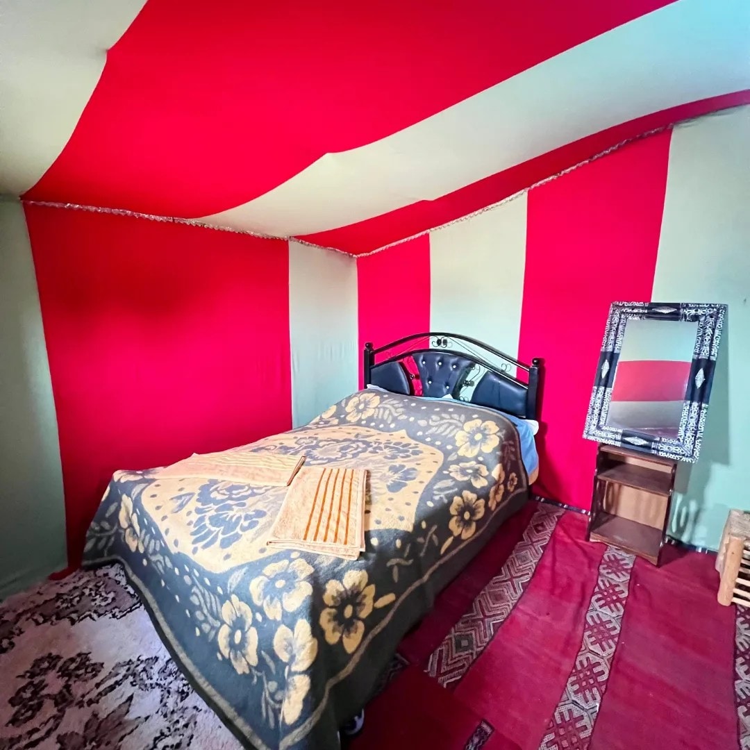 A cozy tent interior features a spacious bed covered with a patterned blanket, situated against vibrant red and green striped walls. A decorative mirror is positioned beside the bed on a wooden nightstand, while textured rugs are laid on the floor, adding warmth to the space.