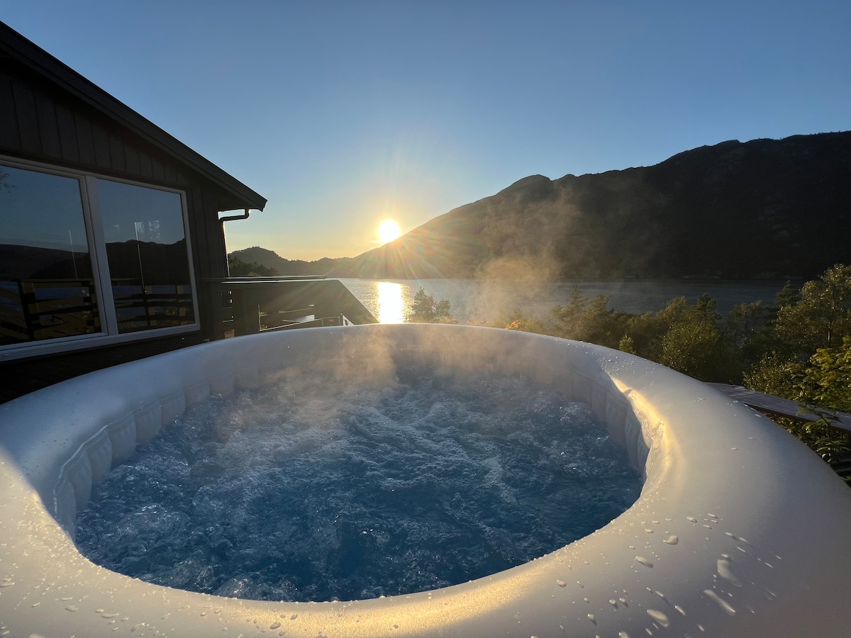 A hot tub filled with steaming water is situated on an outdoor deck, with the sun setting over a mountainous landscape in the background. Soft light reflects off the fjord, creating a serene atmosphere.