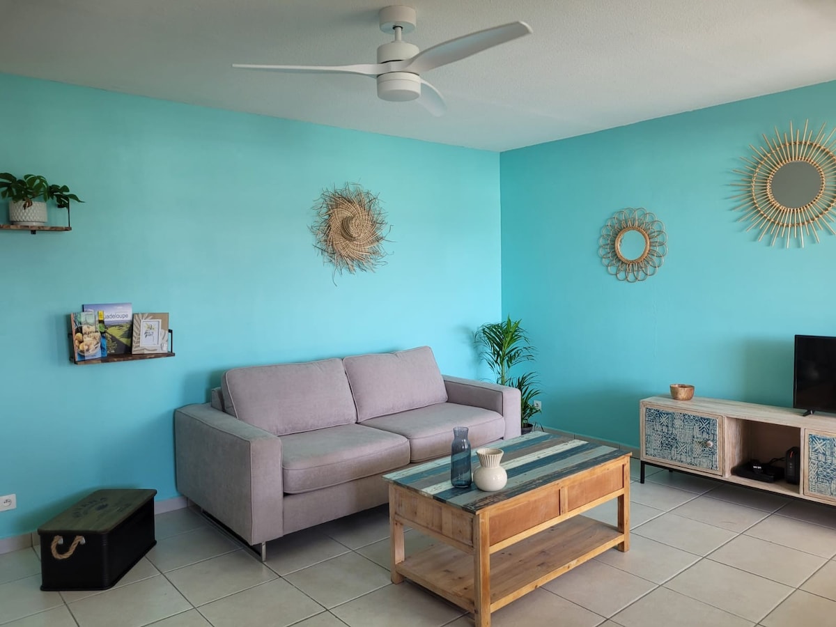 A bright living area features turquoise walls and a comfortable gray sofa. A wooden coffee table with a decorative vase sits in front, alongside a black storage trunk. Natural light is complemented by a ceiling fan, with plants adding a touch of greenery.