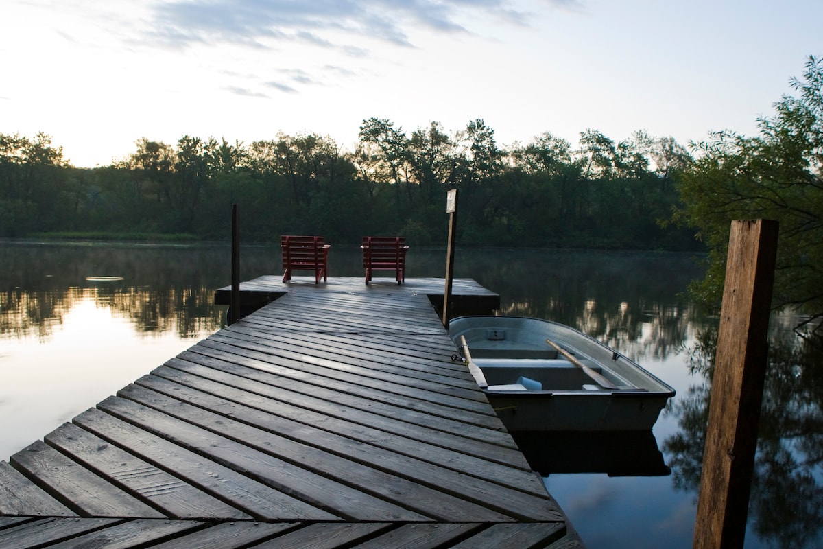A wooden dock extends into a calm river, framed by trees in the background. Two red chairs are situated at the end of the dock, inviting relaxation. A small rowboat is tied alongside the dock, highlighting the tranquil setting.