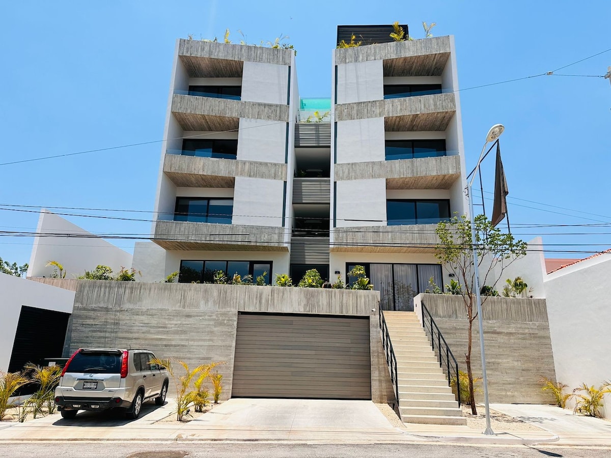 A modern three-story building features a clean design with horizontal concrete and white exterior. Plants are arranged on balconies, providing a natural touch. The entrance is marked by a staircase leading to the main door, and a covered garage is visible on the ground floor.