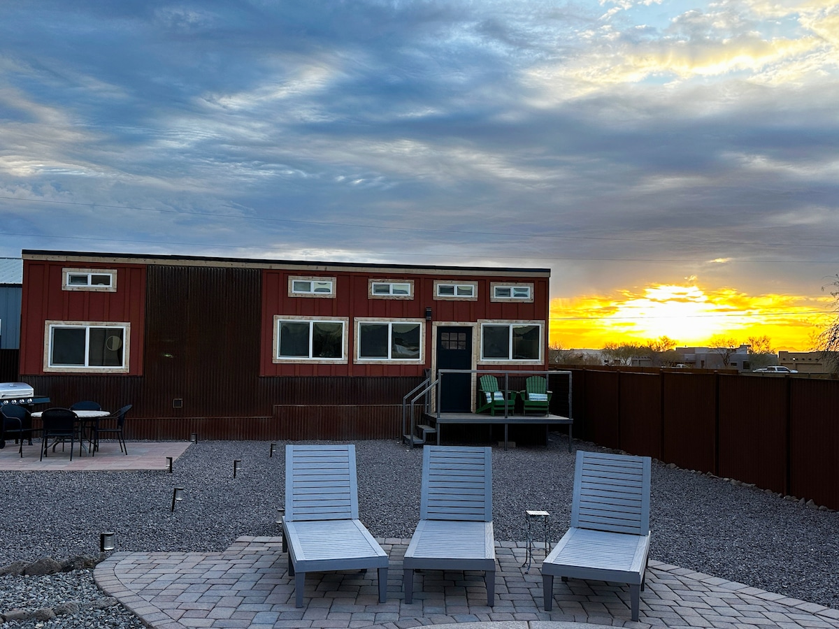 A tiny home is showcased against a vibrant sunset, featuring multiple large windows. In the foreground, three lounge chairs are positioned on stone tiles. A gravel area surrounds the home, enhancing the outdoor space ideal for relaxation.