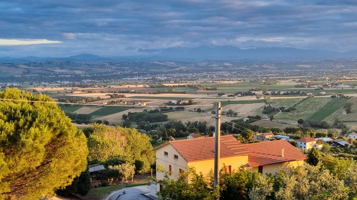 A scenic view is captured, showcasing rolling hills and fields extending into the distance. A two-story house is positioned in the foreground, with a warm light illuminating its exterior. The sky is partly cloudy, creating a serene atmosphere.