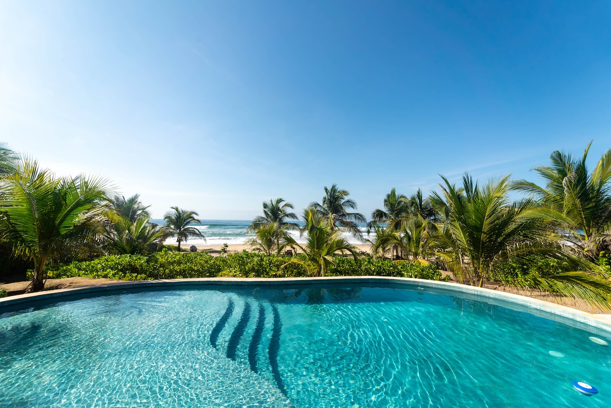 A clear swimming pool reflects the blue sky, bordered by vibrant palm trees. In the background, the ocean waves gently meet the shoreline, creating a serene coastal view.