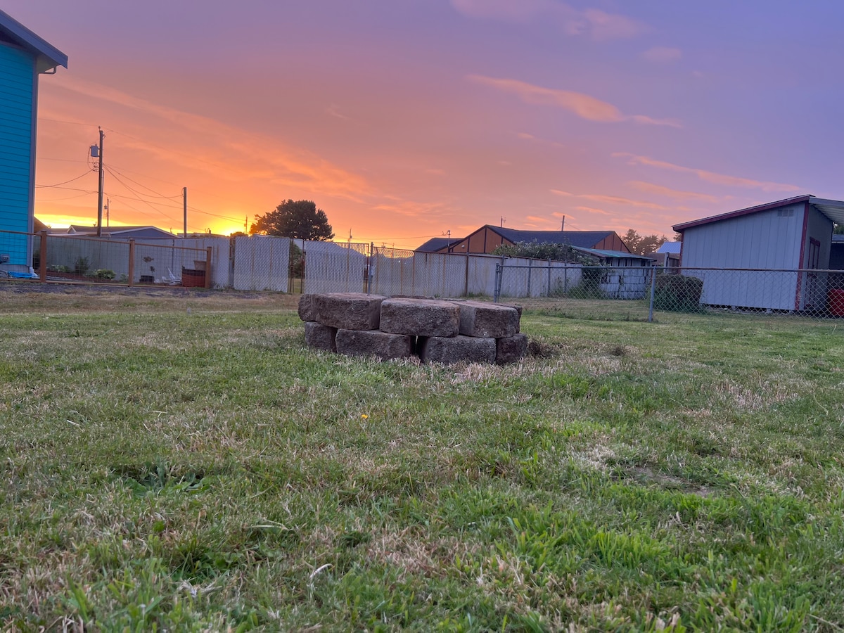 A sunlit backyard features a circular arrangement of stones at its center, surrounded by lush green grass. The background showcases a vibrant sunset with soft hues of pink and orange, creating a tranquil view over the fenced area.