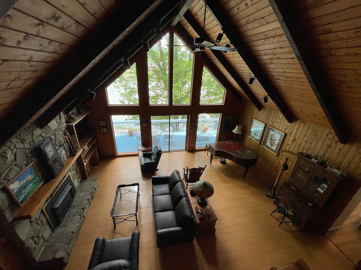 A spacious living area features a high cathedral ceiling supported by wooden beams. Floor-to-ceiling windows provide a view of the lake, while comfortable seating arrangements include a dark leather sofa and armchair. A grand piano is situated near a coffee table, enhancing the inviting layout.