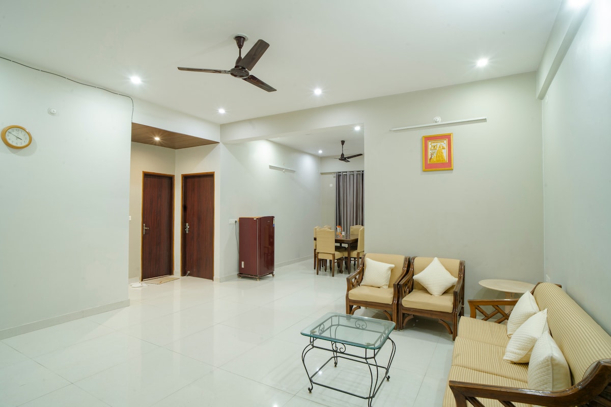A spacious living area is presented with a light-toned tiled floor and neutral walls. Comfortable seating is arranged, featuring a sofa and chairs, complemented by a glass coffee table. A dining area is visible in the background, with a wooden dining table and chairs.