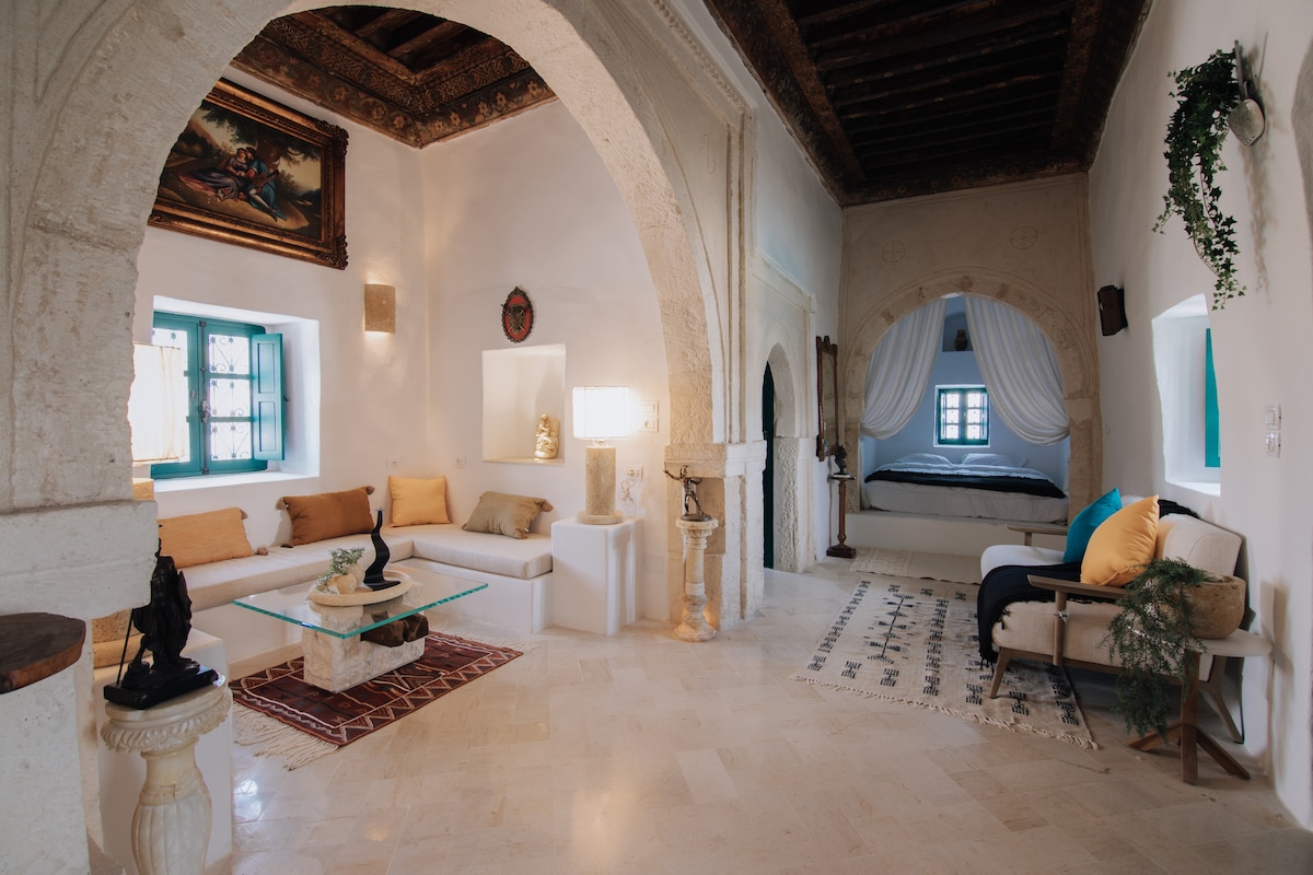 A spacious interior is highlighted by traditional archways, with a low coffee table surrounded by cushioned seating. A dark wood ceiling contrasts with the light-colored walls. A separate sleeping area is visible, featuring an elegant bed draped with sheer fabric. Decorative elements enhance the historic charm.
