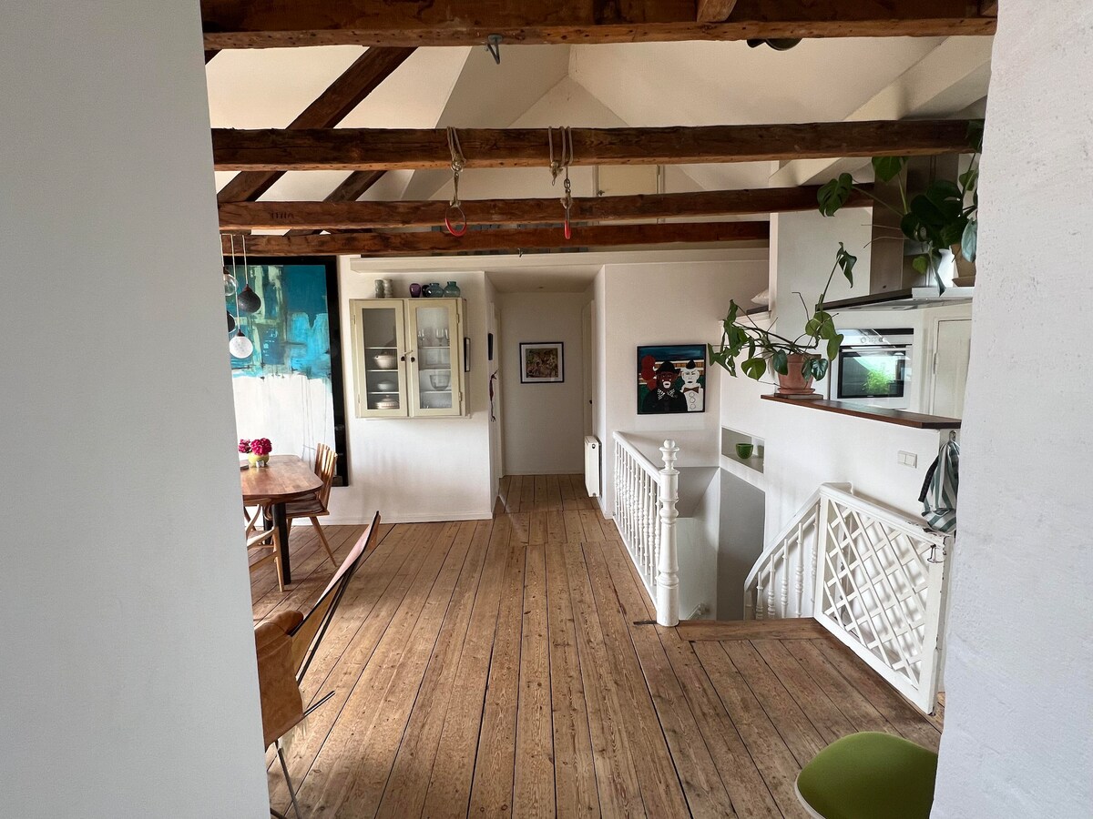 An open and airy living space features wooden beams overhead, complemented by warm-toned flooring. A dining table is positioned nearby with four chairs surrounding it. Plants are arranged in various spots, adding a touch of greenery, while a staircase leads to an upper level.