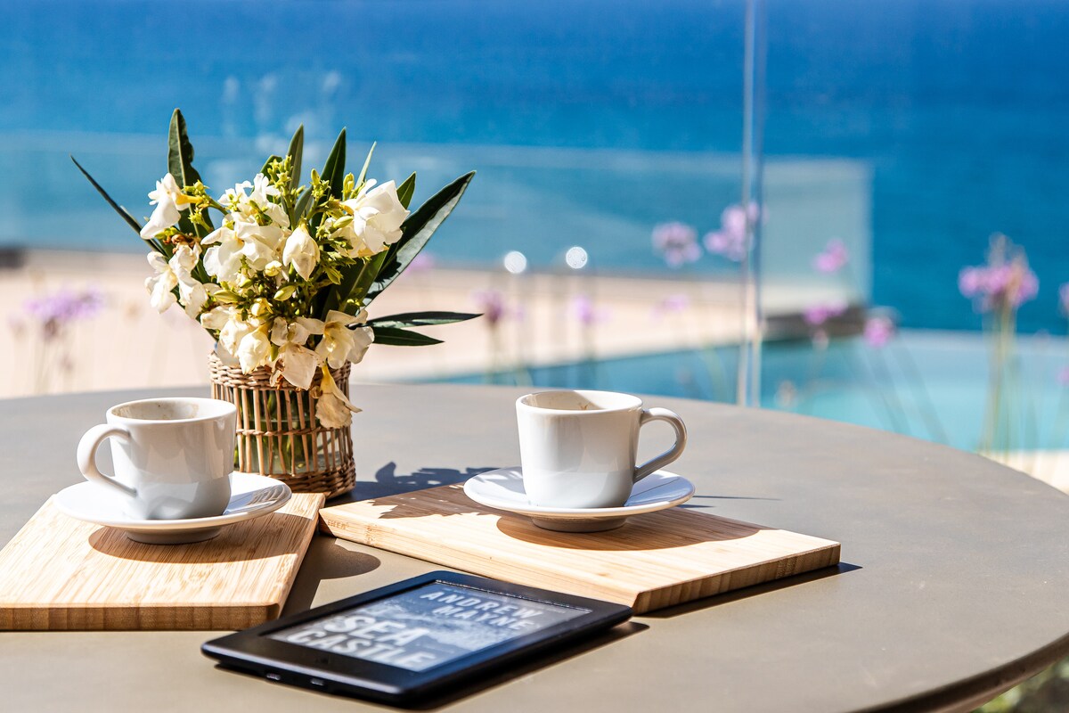 A round table is set for two with white cups and small wooden serving boards. Nearby, a vase of fresh flowers complements the scene. The turquoise sea is visible in the background, enhancing the tranquil setting.