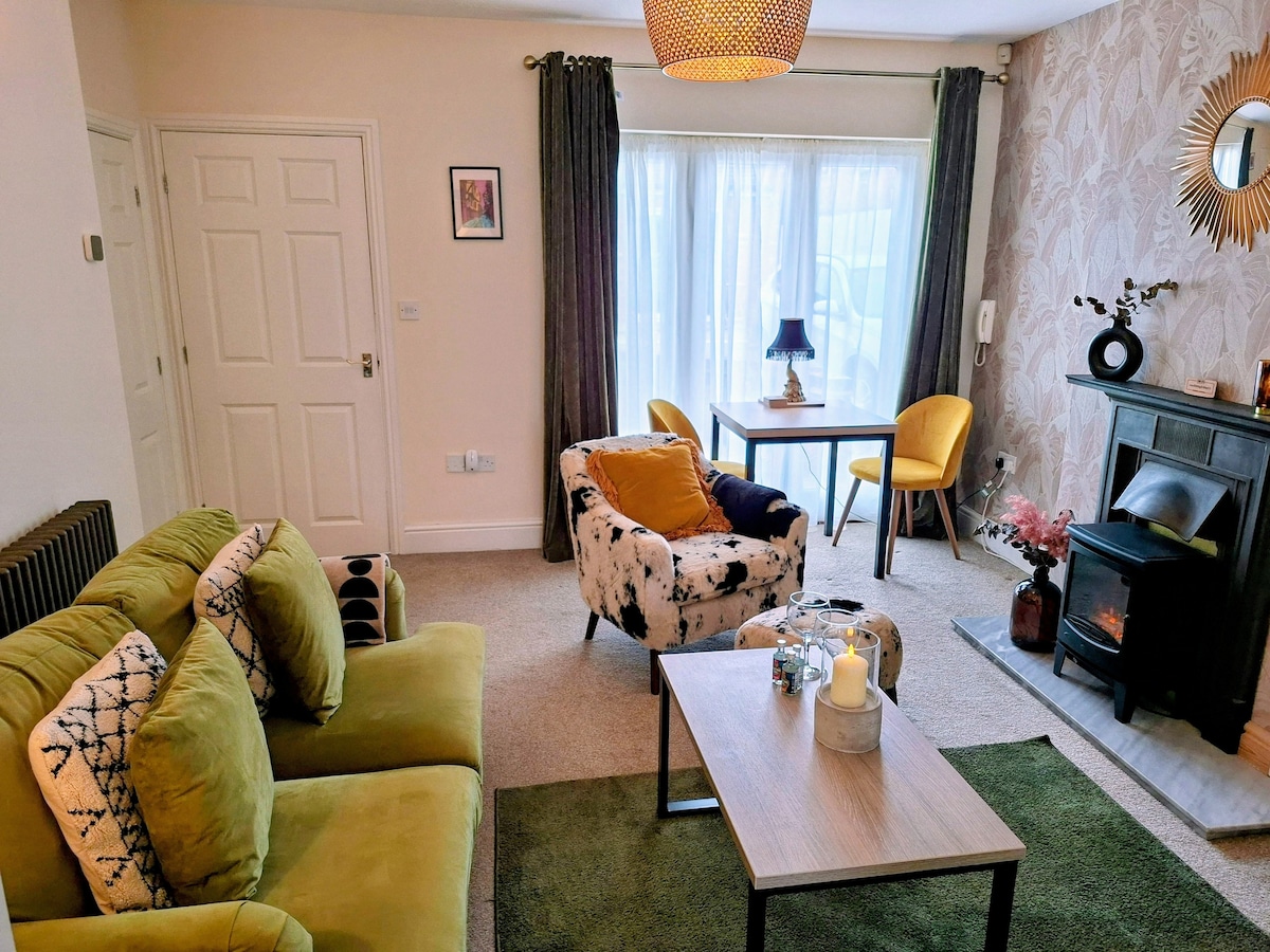 A cozy living area featuring a green sofa with patterned cushions, and a coffee table placed on a light carpet. A dining table with yellow chairs is visible, complemented by light streaming in through large windows covered with sheer curtains. A decorative fireplace adds to the space.