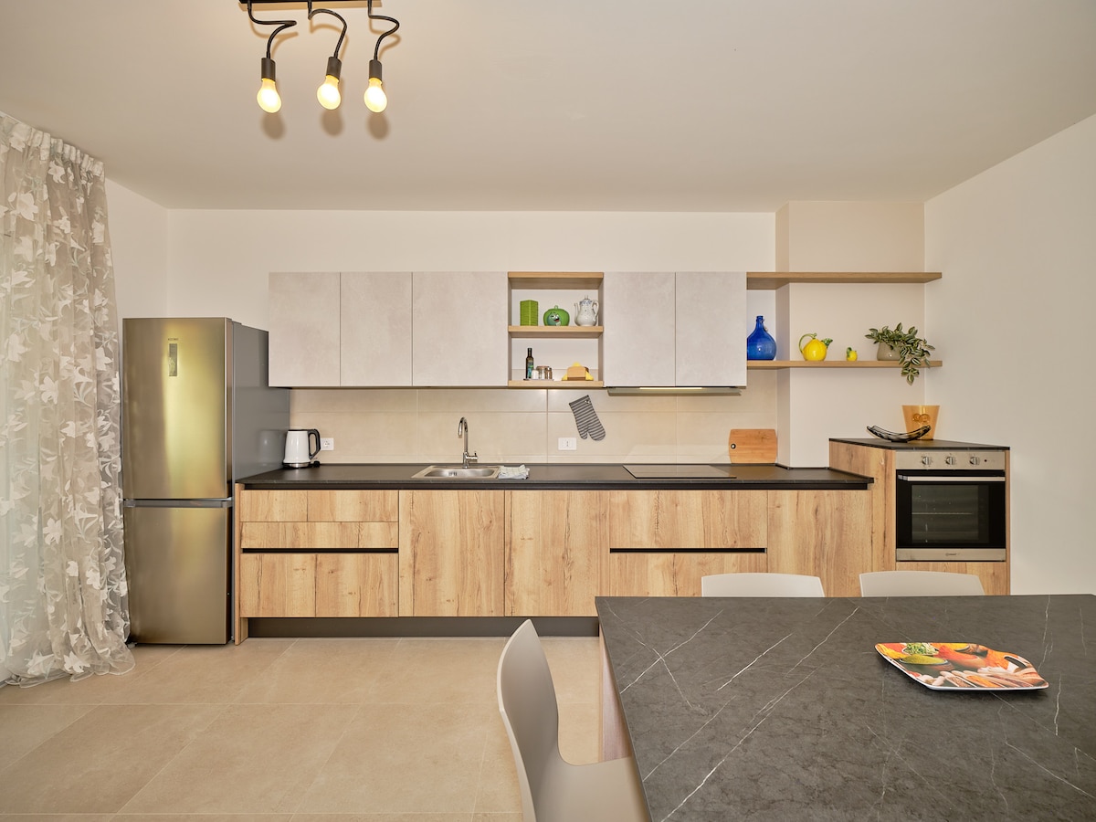 A modern kitchen area is revealed, featuring sleek cabinetry in natural wood tones and a dark countertop. Stainless steel appliances, including a refrigerator, oven, and kettle, are present. A simple dining table with light-colored chairs sits in the foreground, enhancing the functional layout of the space.