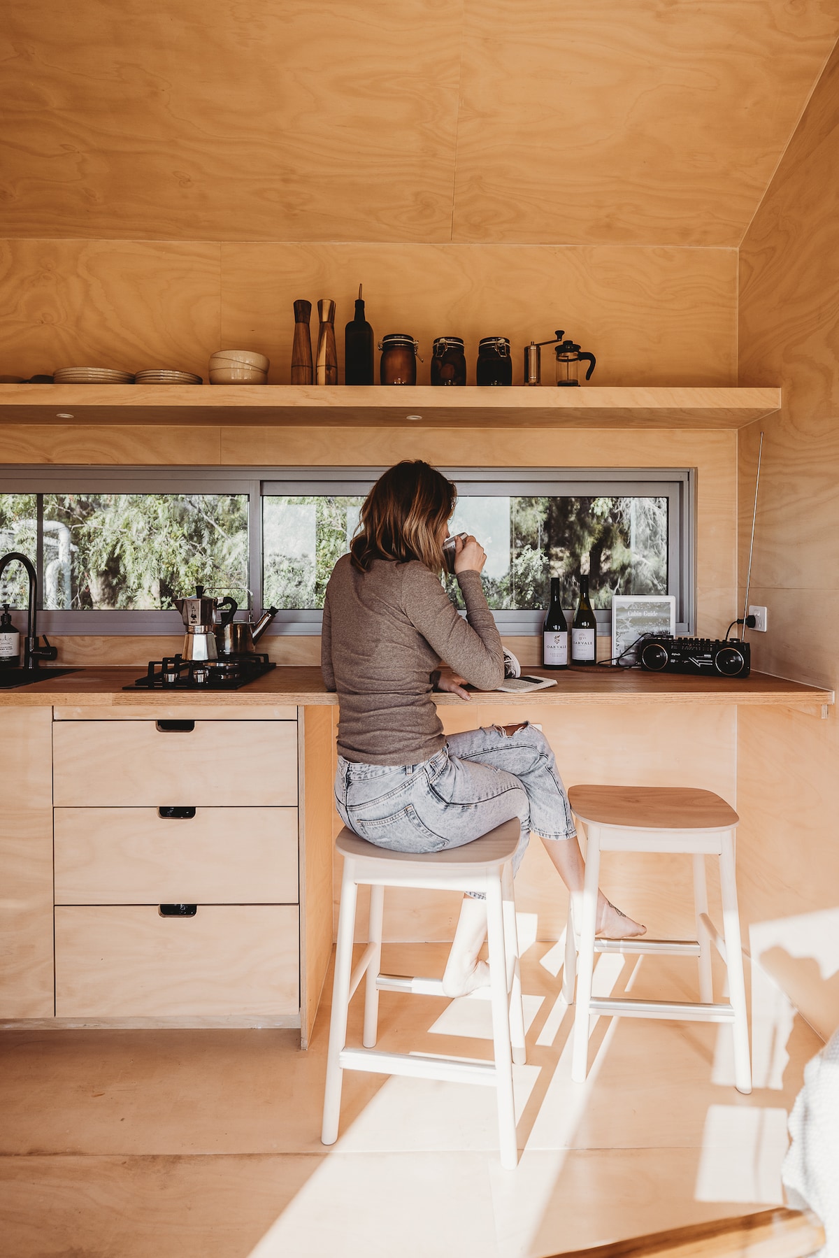A minimalist kitchen area features a wooden counter with essential amenities neatly organized above. A person is seated on a bar stool, enjoying a drink while the sunlight streams through the window, illuminating the space. Wine bottles are displayed on the counter.