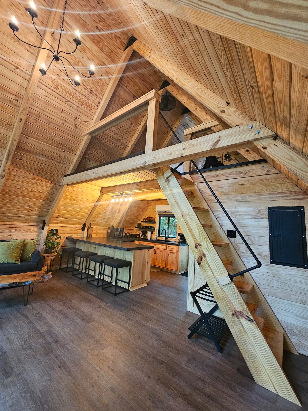 The interior of the A-frame cabin displays natural wood paneling and high ceilings. A spiral staircase leads to the loft area. An open kitchen with wooden cabinetry and bar seating is featured. Soft green cushions are arranged on a sofa, creating a cozy gathering space.