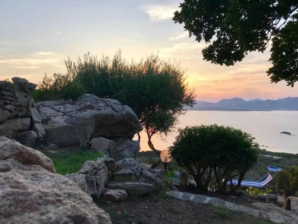 The scene captures a serene sunset over the Gulf of Olbia, with lush greenery and rocky formations in the foreground. Olive trees are visible, and the tranquil waters reflect warm hues from the sky. Mountains create a distant backdrop, enhancing the natural beauty of the area.