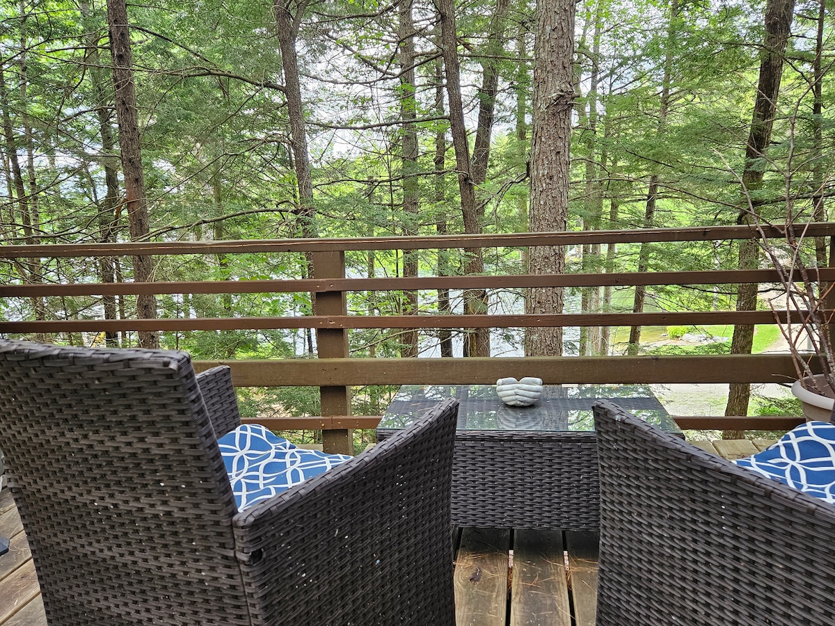 A porch area is furnished with two wicker chairs and a small table, offering a serene view of the surrounding trees and the lake through the wooden railing. Soft blue and white patterned cushions add a touch of comfort to the seating.