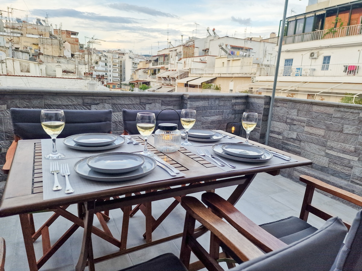An outdoor dining area is featured, showcasing a wooden table set for four with plates, cutlery, and glasses of wine. The surrounding balcony offers views of the urban landscape, with a mixture of buildings and rooftops visible under a cloudy sky.