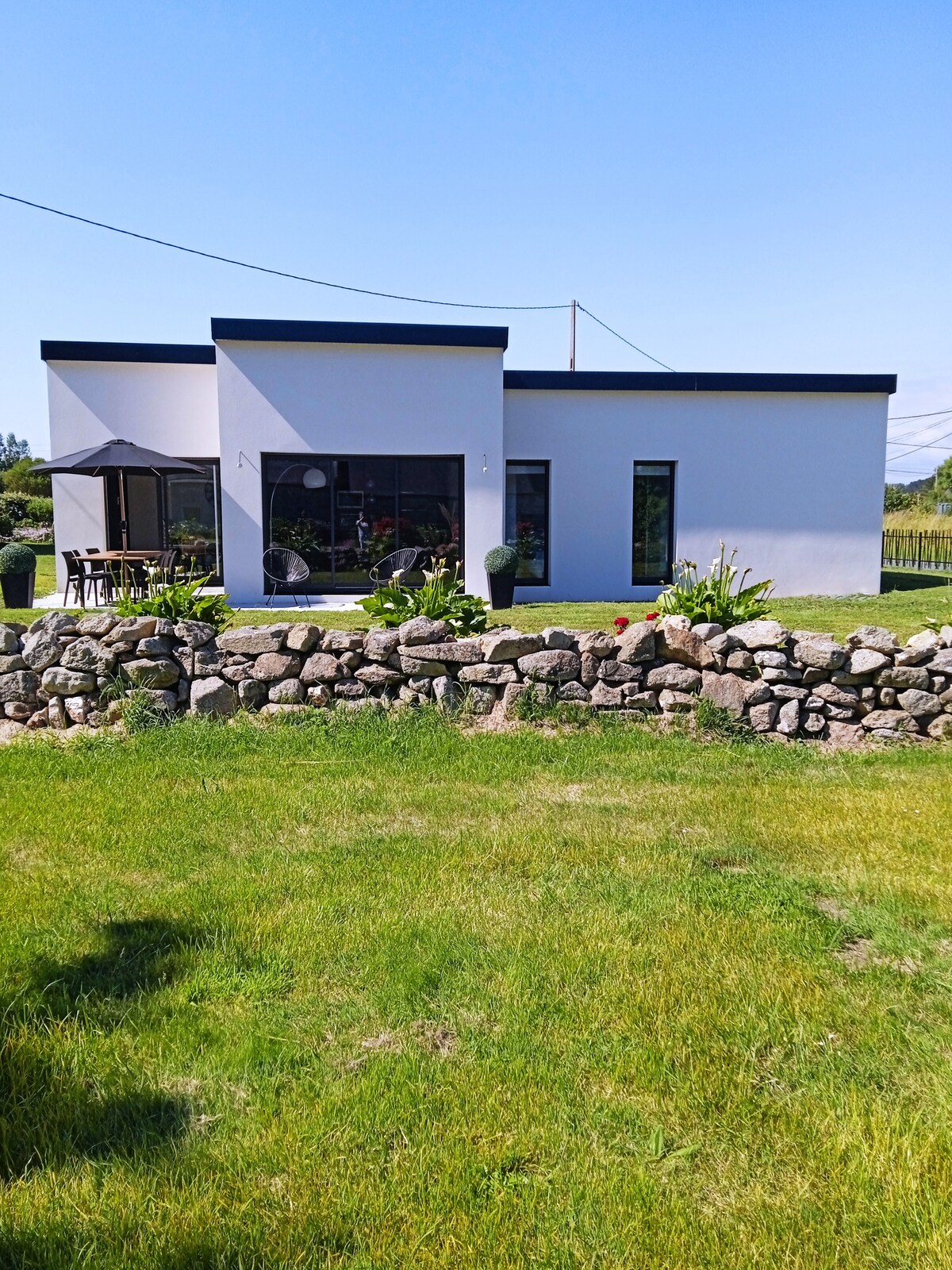 A modern single-story home is set against a bright blue sky, with a well-maintained garden surrounding it. A stone wall borders the grassy area, leading to a patio featuring outdoor furniture. Large windows allow natural light to fill the interior.