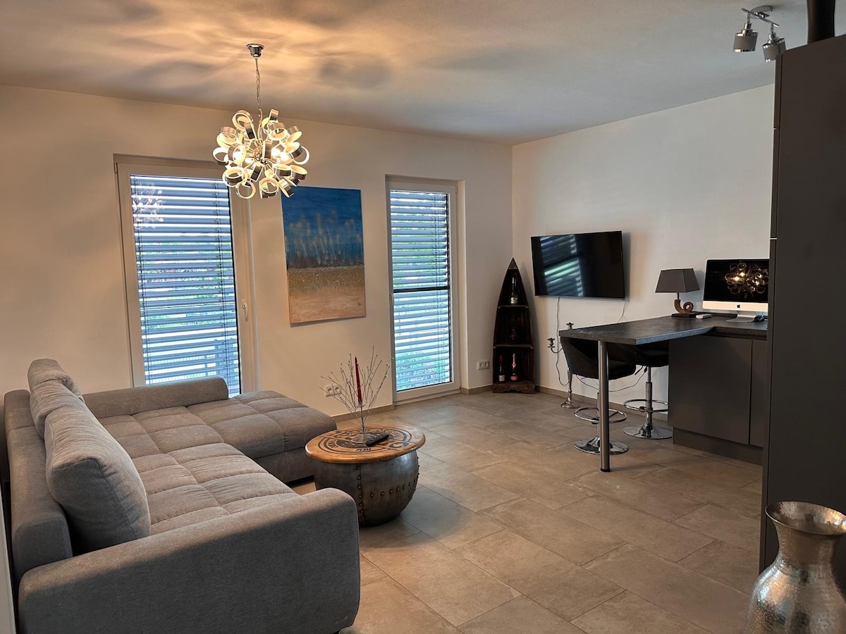 A modern living space features a grey sectional sofa, a wooden coffee table, and a sleek desk with a computer. Natural light is allowed through two windows with horizontal blinds, and a contemporary light fixture hangs from the ceiling. A piece of artwork adds color to the wall.