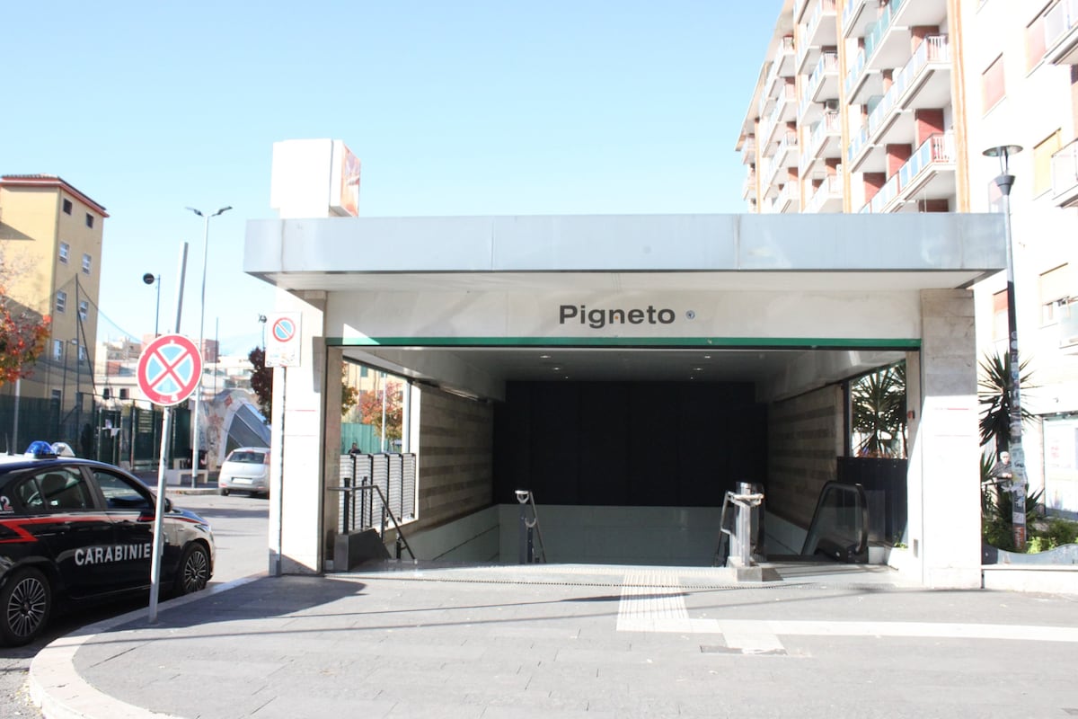 The entrance to the Pigneto metro station is shown, featuring a modern design with a sleek overhang and clear signage. The surrounding area includes plants and nearby buildings, with visible traffic signs and barriers indicating no parking. A police vehicle is parked nearby.