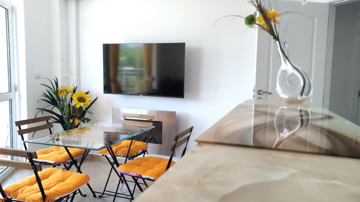 A welcoming living area features a glass dining table surrounded by four brown folding chairs with orange cushions. A large flat-screen TV is mounted on the white wall, while bright yellow sunflowers add a touch of color beside a sleek decorative vase.
