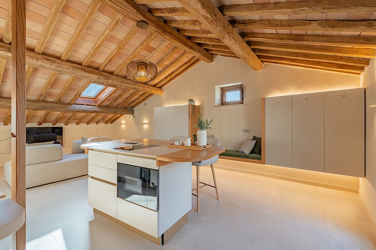 An open-concept kitchen and living area features exposed wooden beams and warm lighting. A central island, with a minimalist design, is surrounded by modern seating. Soft textures and neutral tones create a serene atmosphere. A cozy nook with cushions is visible in the background.