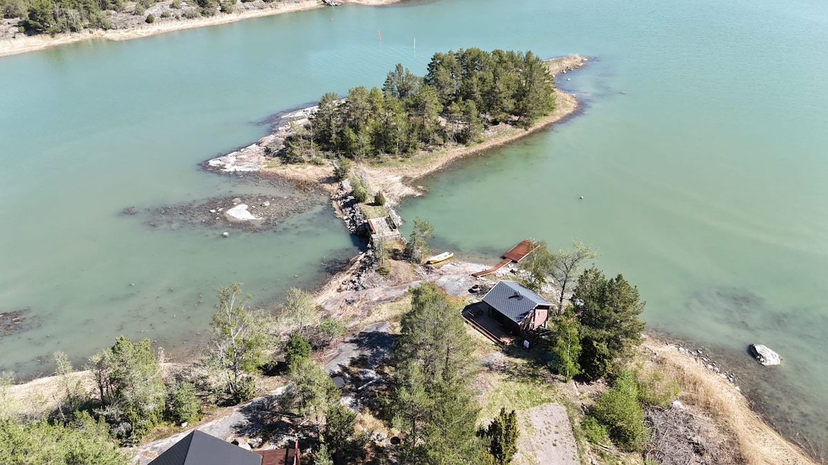 Aerial view of a serene water landscape featuring an island surrounded by calm waters. A small dock extends toward the island, which is adorned with lush greenery. Notable structures include a cabin and a shed, all nestled in the tranquil environment.