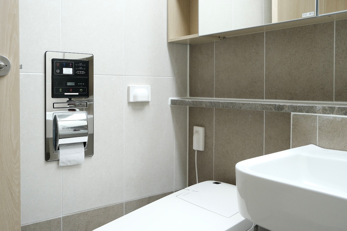 A modern bathroom is depicted, featuring a sleek toilet equipped with a control panel for additional functions. A wall-mounted shelf is visible above the toilet, providing space for toiletries. A clean sink with minimalistic design is positioned nearby, enhancing the contemporary feel.
