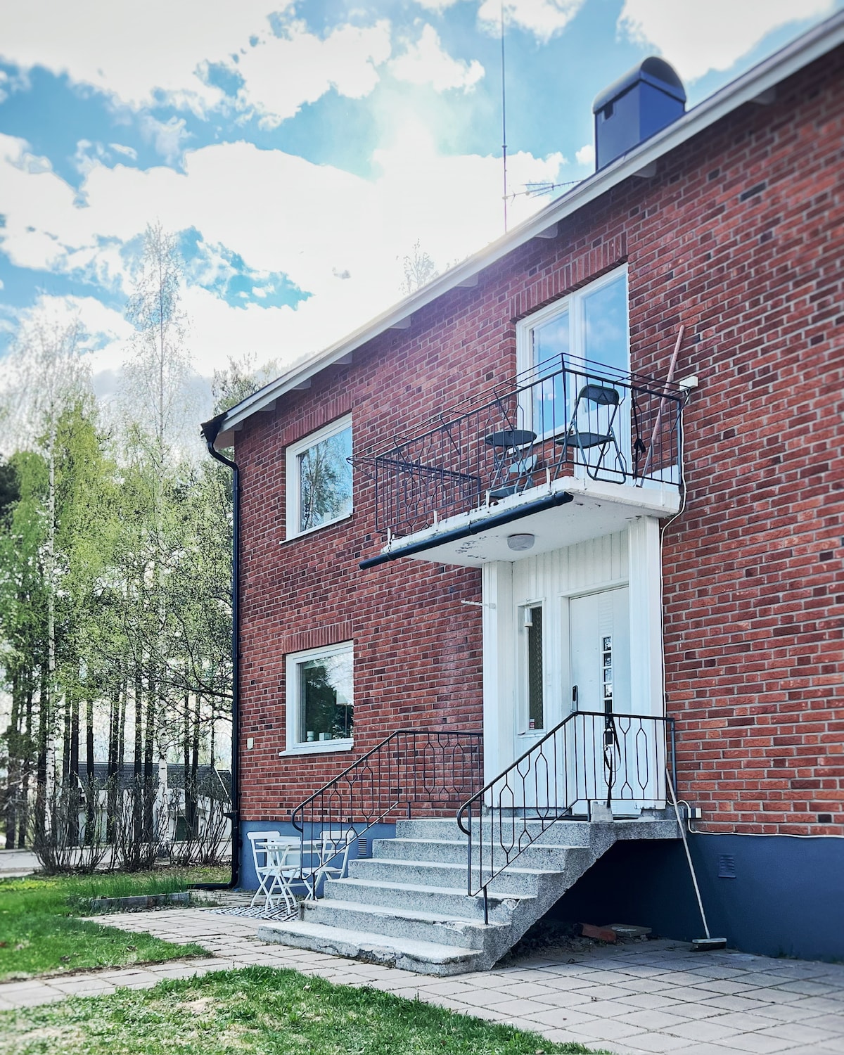 A two-story building features a charming red brick facade and a welcoming entrance. A small balcony with two chairs overlooks the front, and a set of stone steps leads up to the door. Lush greenery surrounds the property, enhancing its inviting appearance.