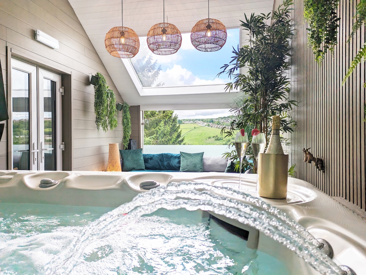 A private hot tub is framed by lush greenery and natural light pouring in through a skylight. Comfortable seating is visible in the background, with rolling hills beyond the glass. The area is designed for relaxation and enjoyment, surrounded by an inviting atmosphere.