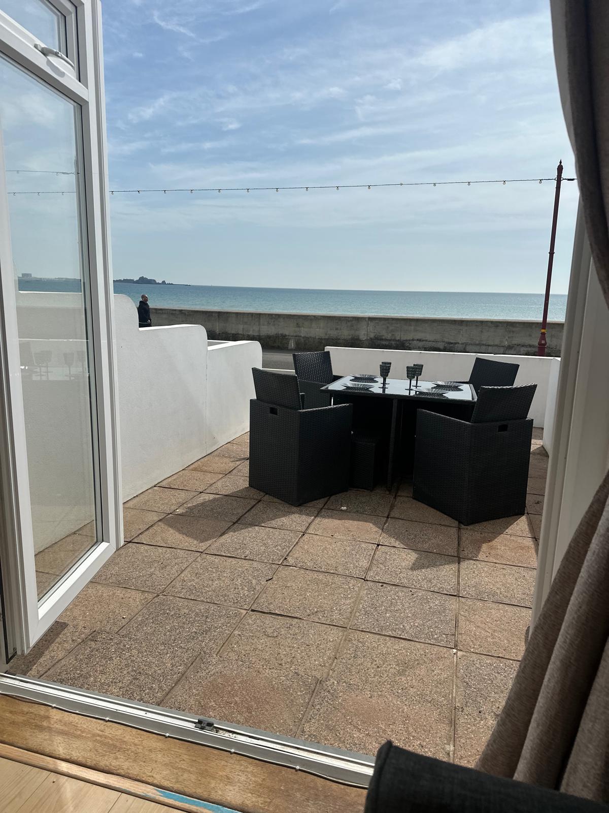 An outdoor patio area is visible, featuring a round table surrounded by five black wicker chairs. The area has a tiled surface and offers an unobstructed view of the sea under a clear sky. A low wall separates the patio from the ocean view.