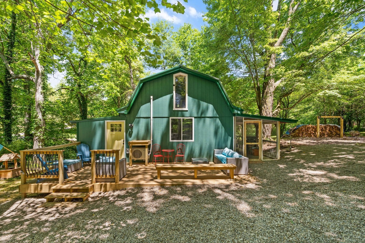 A charming cottage exterior is surrounded by lush greenery, featuring a vibrant green facade. A spacious deck hosts various seating options, including a set of chairs and a sofa. Nearby, a play area is visible, along with a stack of firewood under the trees.