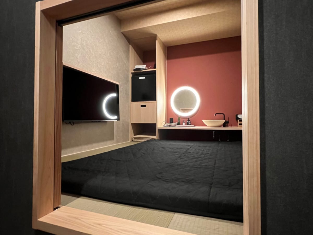 A compact designer room is highlighted by a spacious tatami floor. A dark bedding set is laid out, while a flat-screen TV and a mini-fridge are visible along one wall. A round mirror and sink are elegantly arranged on a counter against a rich red backdrop.