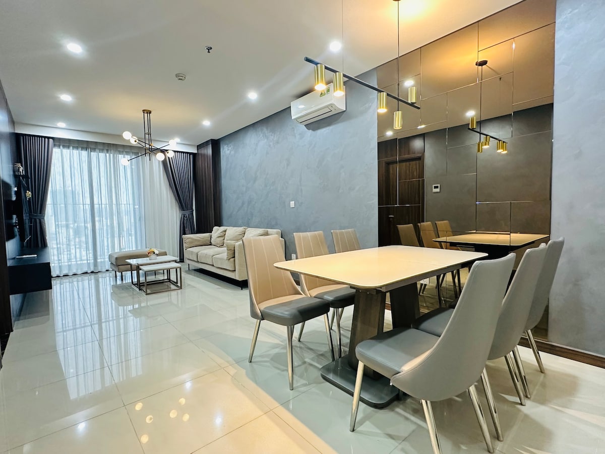 A modern living and dining area features a sleek dining table surrounded by comfortable chairs. A light-colored sofa is positioned against a wall next to large windows that allow natural light to fill the space. Mirrored elements enhance the room's brightness.