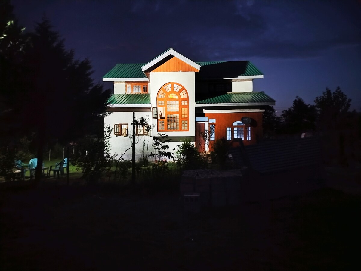 The guest house is illuminated at night, showcasing its distinct architecture with large arched windows. The exterior features contrasting colors with white walls and an orange accent. Nearby green chairs are visible in a garden space, surrounded by trees and foliage.