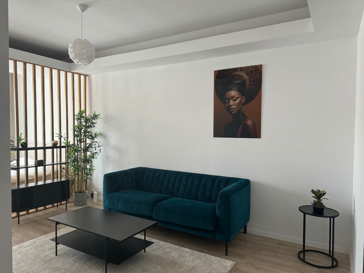 A stylish living area features a vibrant green sofa set against a minimalist white wall. A striking piece of art complements the decor above. A sleek black coffee table occupies the center, surrounded by natural light from a nearby window, with potted plants adding a touch of greenery.