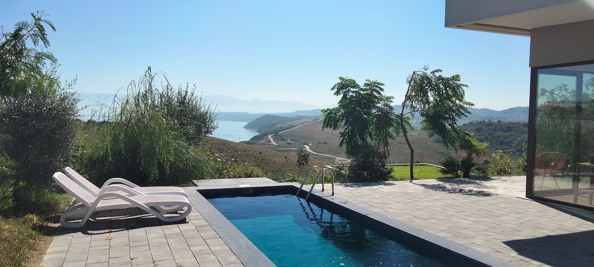 A private pool is set within a spacious stone patio, surrounded by lush greenery. The tranquil landscape features rolling hills and a distant body of water, with sunlight reflecting off the pool's surface under a clear blue sky.