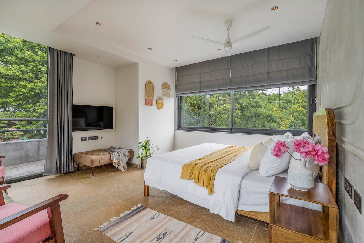 An inviting bedroom is highlighted by large windows that offer views of lush greenery. A spacious bed is adorned with plush linens and decorative pillows. A modern TV and a potted plant add to the room's amenities. Soft lighting creates a calming atmosphere.