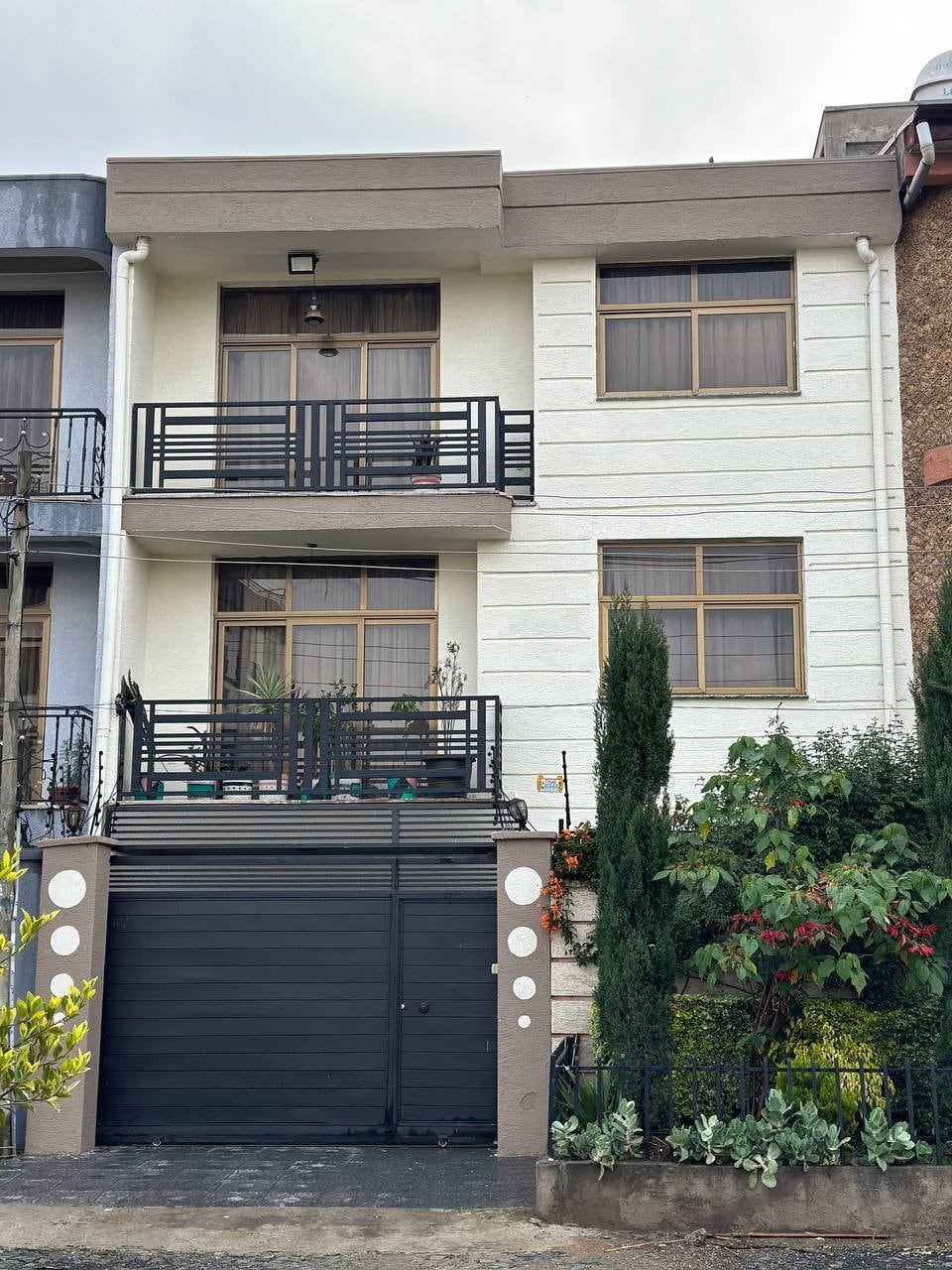 The two-story house features a modern façade with a welcoming entrance. Large windows allow natural light to fill the interior. A secure gate enhances privacy, while landscaped greenery surrounds the property, creating an inviting appeal. The balcony is adorned with potted plants, adding a touch of nature.
