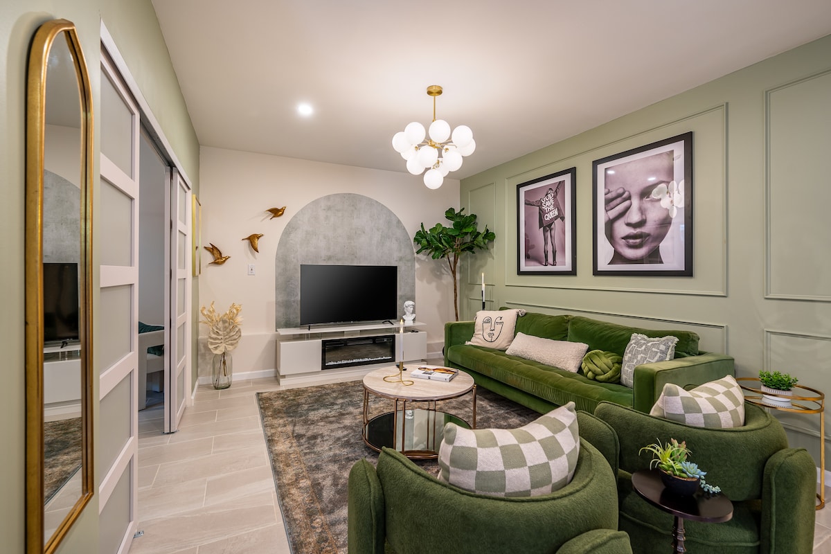 The living area showcases a plush green sofa complemented by stylish armchairs, with framed artwork adorning the walls. A modern chandelier hangs above a round coffee table, while a television is placed against a backdrop of textured paneling. Indoor plants enhance the space's ambiance.