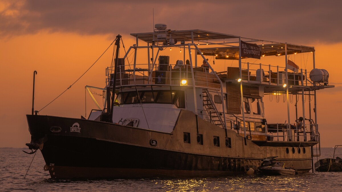 A floating surf hotel is seen anchored in the water during sunset, with soft lighting illuminating the deck and upper levels. The ship features multiple levels, with lounge areas accessible by stairs. A jet ski is stationed alongside, enhancing the nautical theme.