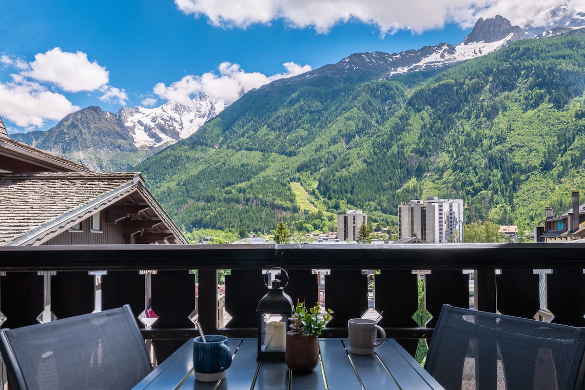 A balcony table with two chairs is set against a backdrop of majestic mountains and green valleys. The table features a small potted plant and a lantern, while a coffee cup and a blue mug are positioned on its surface. Breathtaking views of the surrounding landscape are visible.