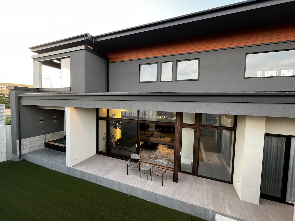 The exterior view of the villa showcases a modern design with large glass windows, providing ample natural light. A spacious patio area is visible, featuring seating arrangements. The well-maintained lawn complements the elegant structure, creating an inviting outdoor space.