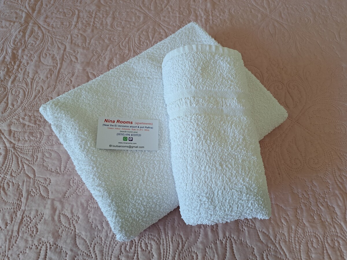 Fresh white towels are neatly arranged on a light pink quilted surface. A small card with contact details is placed beside the towels, providing clear identification of the accommodation.