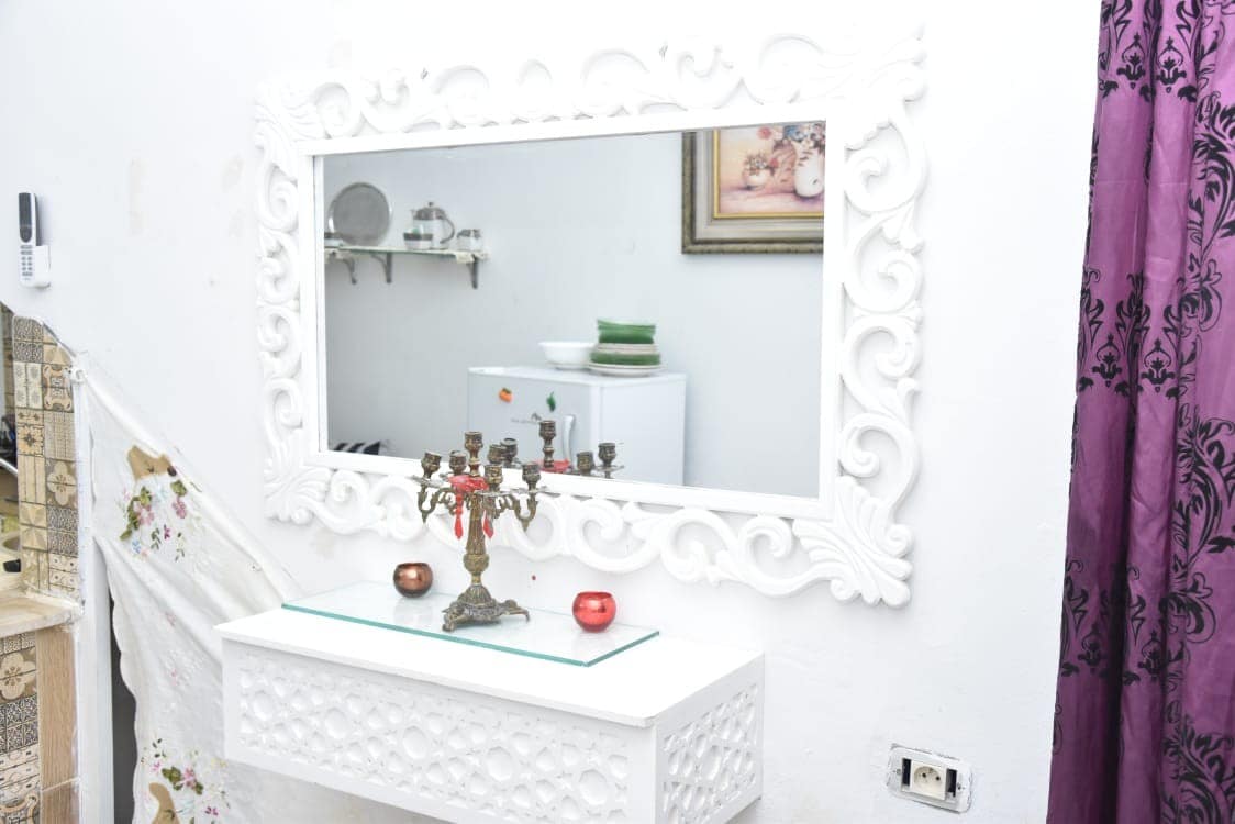 A white decorative mirror is positioned above a sleek console table, reflecting the surroundings. Soft purple curtains frame the space, while a small decorative candelabrum and candles are placed on the table. A white cabinet and wall art are visible in the background.