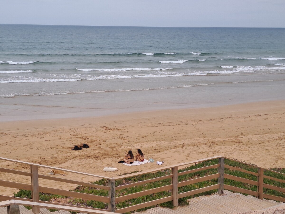 A sandy beach is visible with gentle waves lapping at the shore. Two individuals are seen sitting on a light-colored blanket, enjoying the view. Wooden railings frame the scene, leading toward the beach, while patches of grass add a natural touch to the setting.